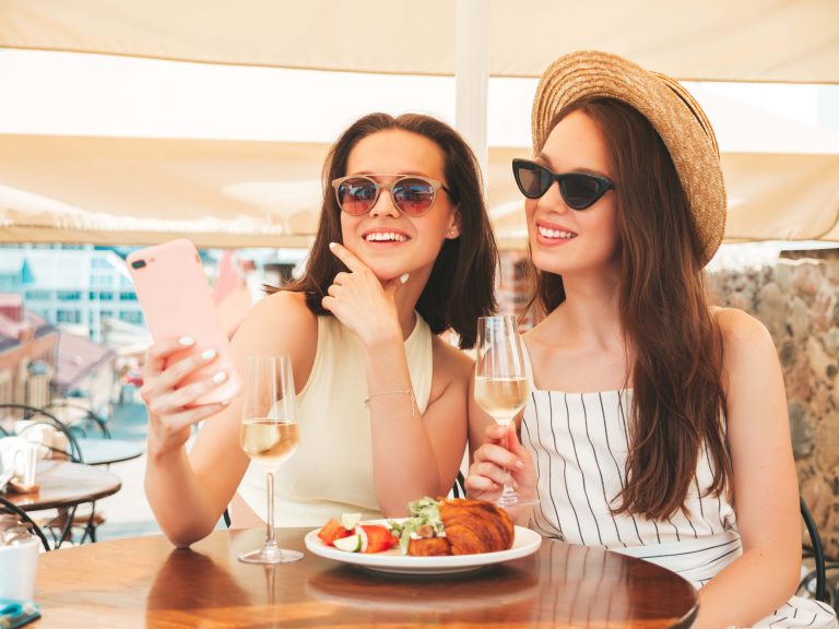 two young beautiful smiling hipster female summer clothescarefree women posing veranda cafe streetpositive models drinking white wine hatlooking smartphone screen using apps scaled